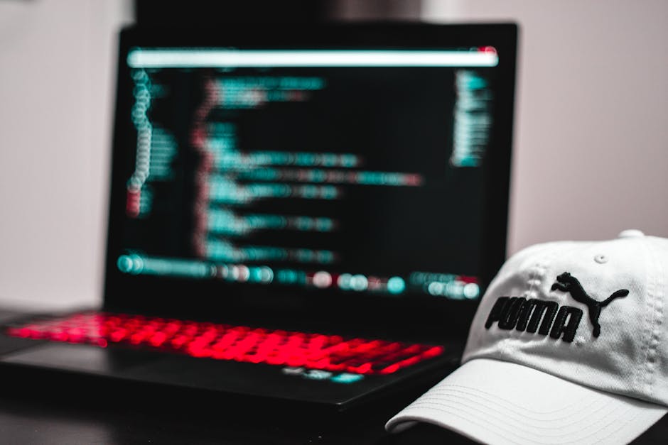 A person operating a laptop computer on a dark surface, with their hands visible pressing keys. The laptop screen displays lines of colorful code or programming scripts on a black background, indicati