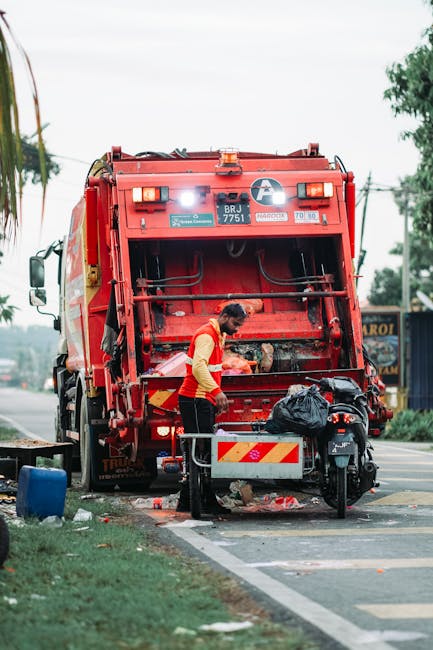 A large pile of mixed household waste and recyclable rubbish situated on a paved sidewalk in an urban setting. The collection includes open cardboard boxes, crumpled paper and packaging, plastic bags,