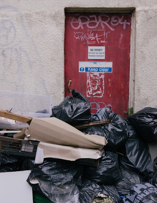 A pair of large black garbage bags and a cardboard box filled with waste materials are positioned on a pavement next to a black metal fence, with lush leafy bushes behind the fence. The bags appear to
